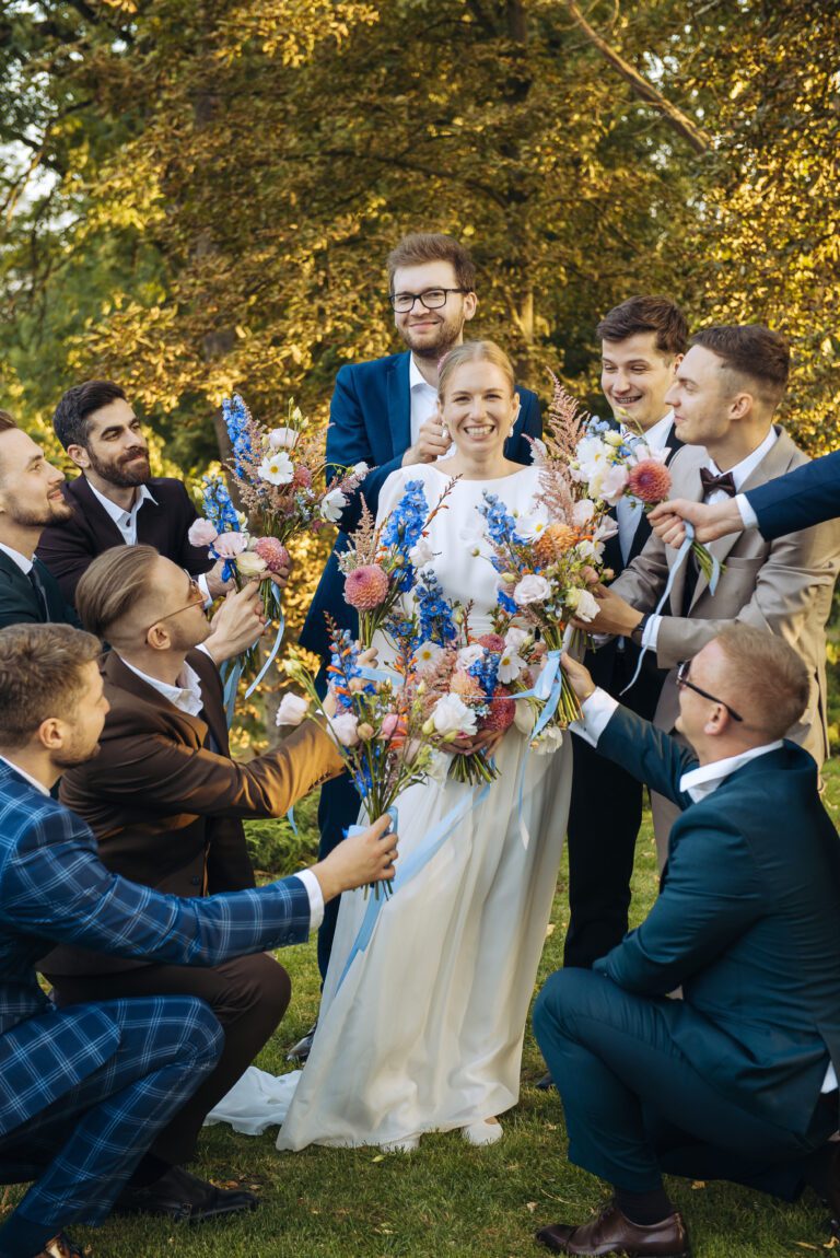 Panna młoda otoczona przez koleg&oacute;w, kt&oacute;rzy trzymają kwiaty i klękają przed panną młodą.