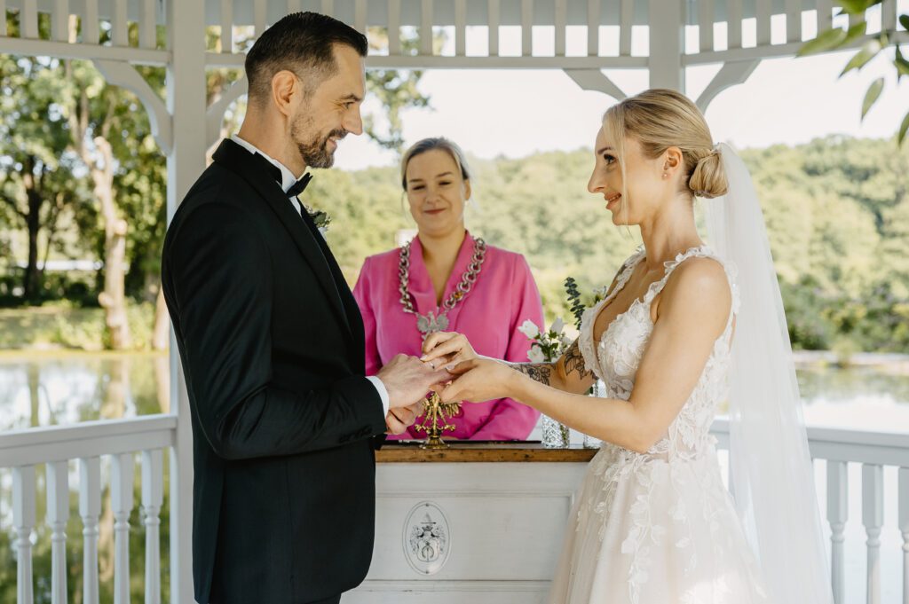 Panna młoda zakłąda obroczkę panu młodemu w altanie w obecności urzędnika.