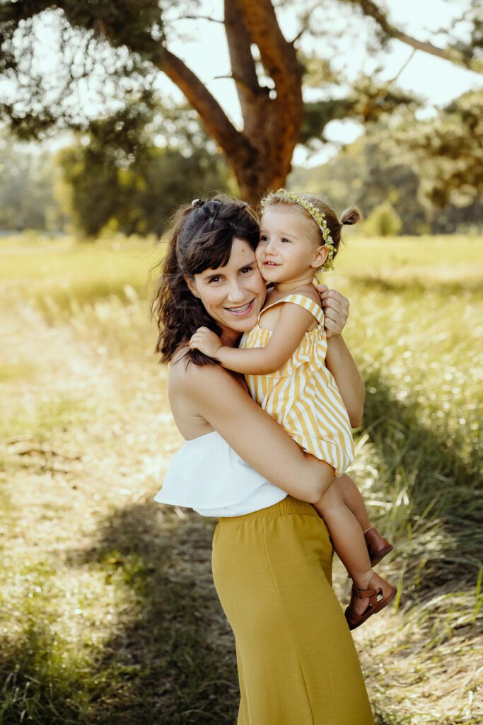 DSC_5210 Mama przyytula się z córeczką. Trzyma ją na rękach. Mam patrzy w obiektyw. Jest lato. Mają naturalną lnianą odzież.
