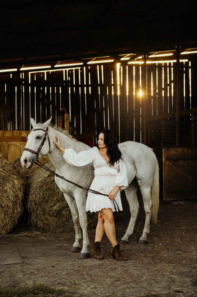 portret kobiety z koniem w starej stodole, gdzie przez deski prześwituje słońce