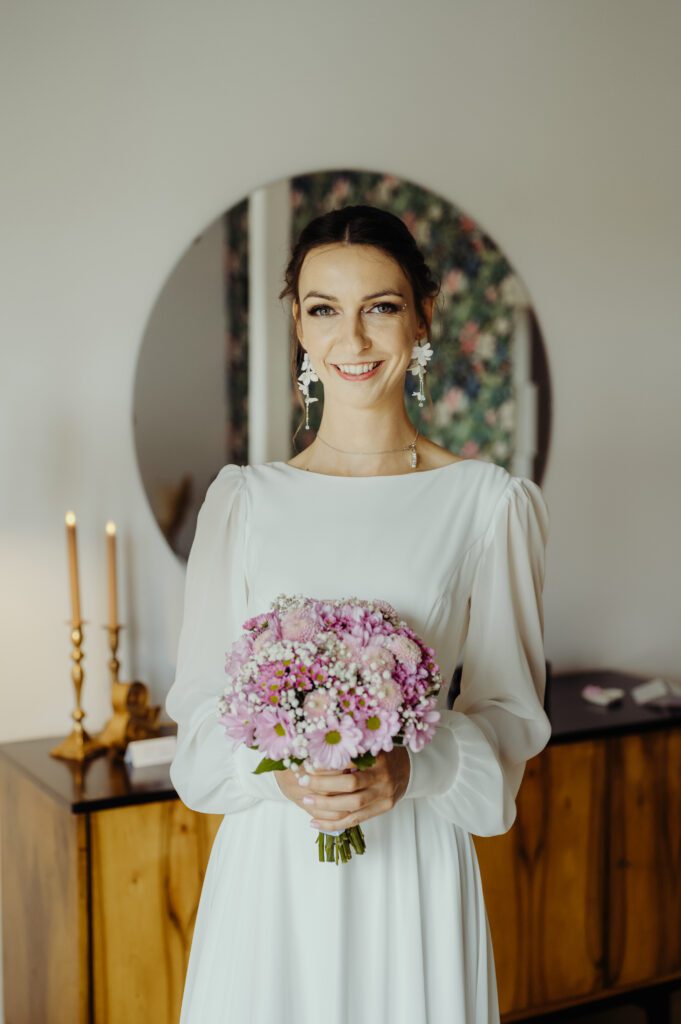 300525_0167 panna młoda Marta w sukni ślubnej, trzyma bukiet ślubny i partrzy prosto w aparat uśmiechając się