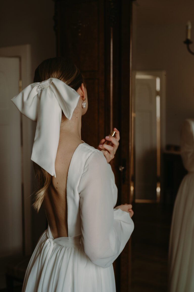 Pann młoda w sukni ślubnej przegląda się w lustrze i skrapia perfumami. Widać tył sukienki, gołe plecy oraz dużą kokardę we włosach.