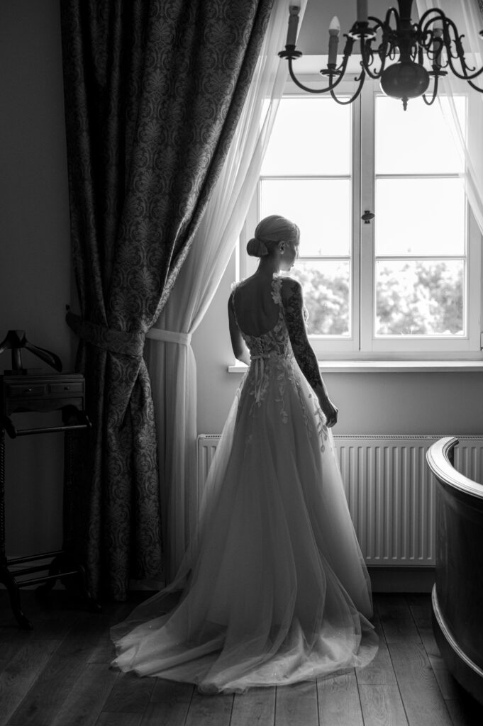Nastrojowe czarno-białe zdjęcie panny młodej w sukience, spoglądającej przez okno.
