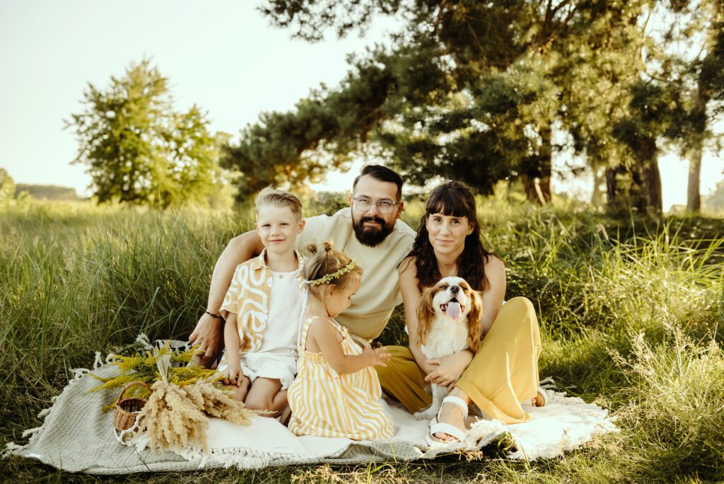 DSC_1069 Rodzinka z pl,em siedzą na kocu. Jest pieknyy letni dzień. Na kocu leża świeżo zebrane kwiaty i trawy. Mam trzyma psa. Tata z synem patrzą w obietyw. Mała dziewczunka bawi się.