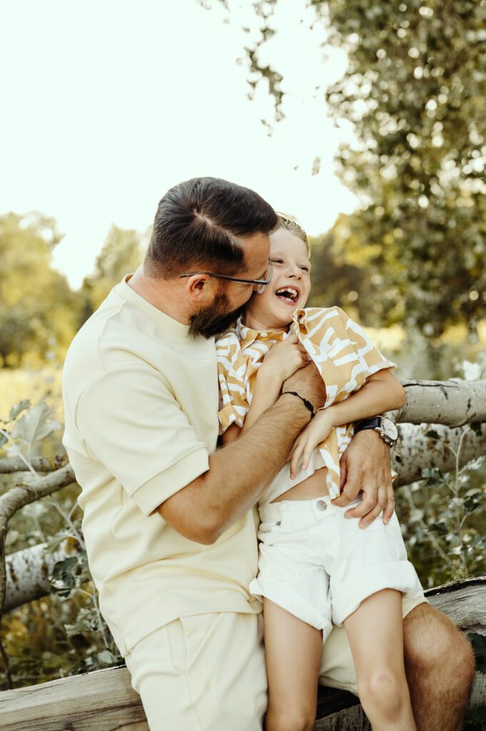 DSC_5379 Tata wygłupia się z synem. Sun się śmieje. Tata go łaskocze. Oboje siędzą na powalonym pniu drzewa. Jest piękny letni dzień.